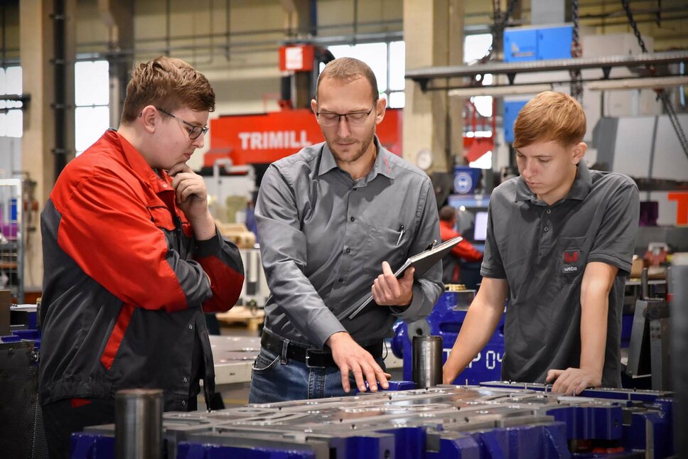 Das Bild zeigt den Lehrlingsausbilder, der zwei Lehrlingen in der Werkzeugbautechnik in einer Produktionsumgebung ein Umformwerkzeug erklärt. Er zeigt auf bestimmte Teile des Werkzeugs und erklärt die Funktionen und den Zweck jeder Komponente. Die Lehrlinge hören aufmerksam zu und stellen Fragen, während sie lernen. Die Szene vermittelt den Eindruck von Wissensaustausch und praktischer Schulung in einem professionellen Arbeitsumfeld.