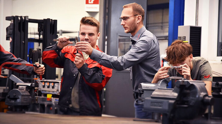 Ein Lehrling in der Werkzeugbautechnik, konzentriert beim Messen eines Stahlklotzes mit einem Messschieber, während der Ausbilder neben ihm steht und etwas erklärt und zeigt. Daneben spannt ein weiterer Lehrling aufmerksam einen Stahlklotz in einen Spannbock, was die praktische und lehrreiche Umgebung der Ausbildung verdeutlicht.