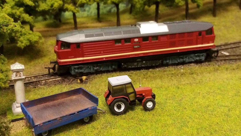 Detailaufnahme einer Modelleisenbahn mit roter Lokomotive, Traktor und Anhänger in einer gestalteten Landschaft.