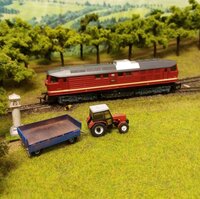 Detailaufnahme einer Modelleisenbahn mit roter Lokomotive, Traktor und Anhänger in einer gestalteten Landschaft.