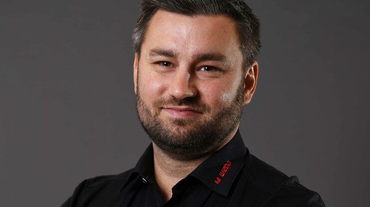 Alexander smiles confidently at the camera, wearing a black shirt with a red weba logo on the collar, posing in front of a neutral grey background.