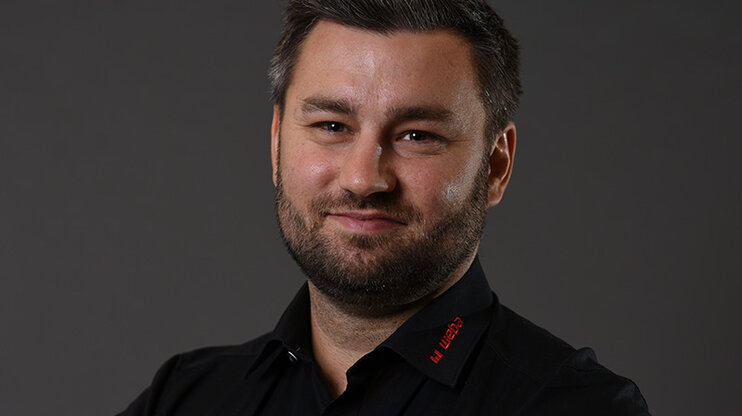 Alexander se prezentuje přátelsky a profesionálně: černá košile s červeným logem weba na šedém pozadí, usmívá se přímo do kamery.