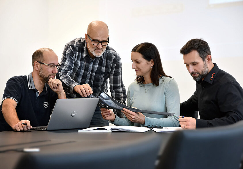 Vier Personen sitzen an einem Besprechungstisch: Eine Person arbeitet an einem Laptop, eine andere betrachtet aufmerksam Unterlagen, während zwei weitere Personen sich angeregt über ein Stanzteil unterhalten. Die Szene zeigt eine konzentrierte und engagierte Arbeitsatmosphäre.